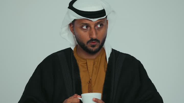 التلذذ والاستمتاع بتناول المشروب الساخن في الجو الشتوي البارد، الشعور بالصقيع والقشعريرة والتجمد، ارتداء الملابس الدافئة في فصل الشتاء، إيماءات تدل على البرد الشديد، تصوير مقرب لرجل عربي خليجي اماراتي يرتدي الفروة و والغترة يقوم بشرب كوب من القهوة، خلفية بيضاء
