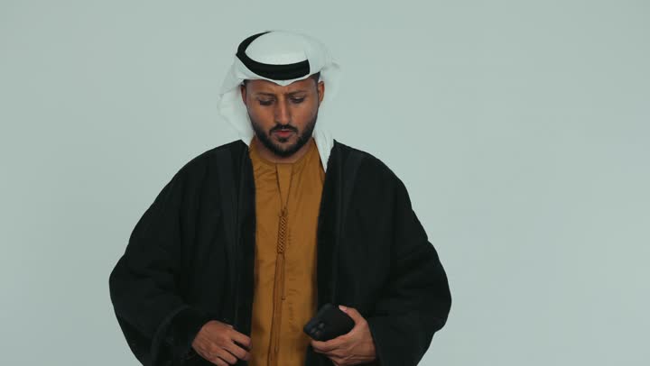 التقاط صورة سيلفي تذكارية باستخدام الجوال، ارتداء الملابس المناسبة لفصل الشتاء في الأجواء الشتوية الباردة، الشعور بالصقيع والقشعريرة والتجمد، إيماءات تدل على البرد الشديد، رجل عربي خليجي اماراتي يرتدي الفروة و والغترة يمسك بيده الهاتف المحمول، خلفية بيضاء