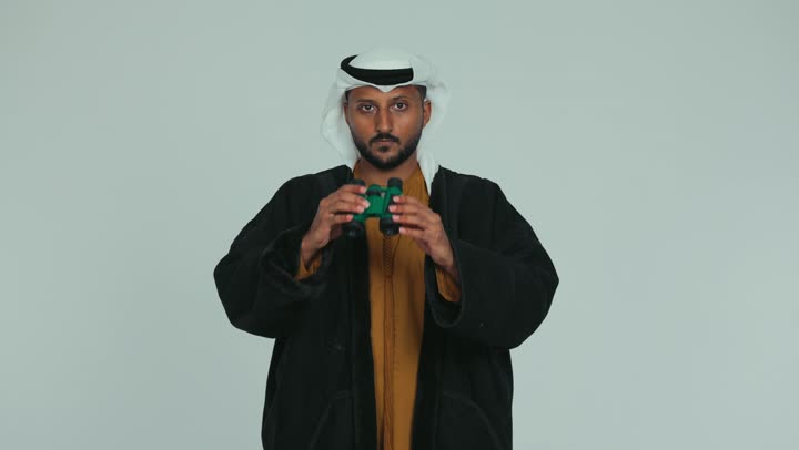 مراقبة ومشاهدة الأشياء البعيدة، الرؤية المستقبلية وتحديد الأهداف والطموحات، رجل عربي خليجي اماراتي يرتدي الكندورة والغترة يمسك بيده  منظار ثنائي العينين وينظر من خلاله بإيماءات السرور والسعادة، خلفية بيضاء