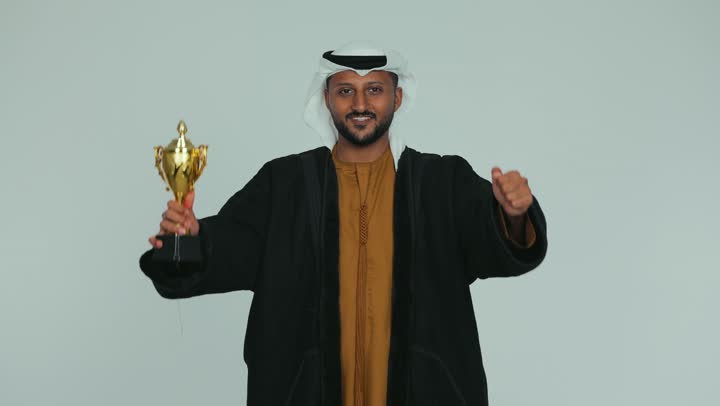 أنشطة ترفيهية ممتعة، النظر إلى الكاميرا بإيماءات الفرح والسعادة لفوز الفريق المفضل، تشجيع الأنديه والمنتخبات والفرق، رجل عربي خليجي اماراتي مبتسم يرتدي الكندورة والغترة يمسك بيده الكأس الذهبي، رفع اليدين بتعابير الانتصار، خلفية بيضاء