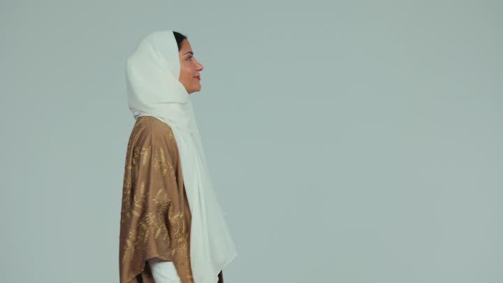 ايماءات إلقاء التحية والسلام، تعابير الترحيب والاستقبال، محاولة لمس شيء ما، تصوير من الجانب لإمرأة عربية خليجية اماراتية محجبة ترتدي عباءة تمشي وتقوم برفع يدها للأعلى بتعابير السعادة والسرور، خلفية بيضاء