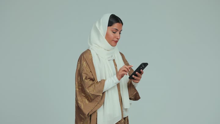 Taking souvenir selfies, taking advantage of modern technological devices in free time, a veiled Emirati Gulf Arab woman wearing an abaya using a mobile phone with gestures of happiness and pleasure, chatting and communicating with family and friends via mobile, white background