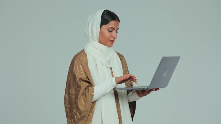Merging technology in practical life, chatting and communicating with family and friends through the modern technical system, remote work and browsing the Internet using a computer, a veiled Emirati Gulf Arab woman wearing a mantle holding her computer and holding a meeting online, white background