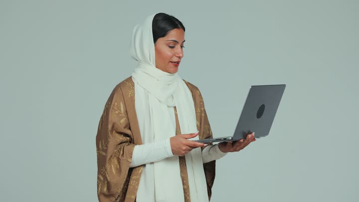 Remote work and browsing the Internet using the computer, integrating technology into practical life, chatting and communicating with family and friends through the modern technical system, a veiled Arab Gulf woman who wears a mantle holding her computer and holds a online meeting, white background