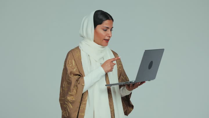 Merging technology in practical life, chatting and communicating with family and friends through the modern technical system, remote work and browsing the Internet using a computer, a veiled Emirati Gulf Arab woman wearing a mantle holding her computer and holding a meeting online, white background