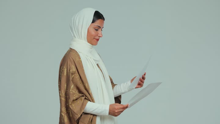 Proofreading written notes, preparing work reports, side view of a veiled Emirati Gulf Arab woman wearing an abaya holding white papers, reviewing and following up on plans and projects, exchanging experiences and skills, white background
