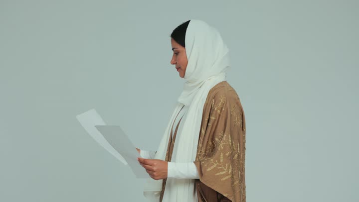 التدقيق في الملاحظات المكتوبة، اعداد التقارير الخاصة بالعمل، تصوير من الجانب لامرأة عربية خليجية اماراتية محجبة ترتدي العباءة تحمل بيدها أوراق بيضاء، مراجعة ومتابعة الخطط والمشاريع، تبادل الخبرات والمهارات، خلفية بيضاء