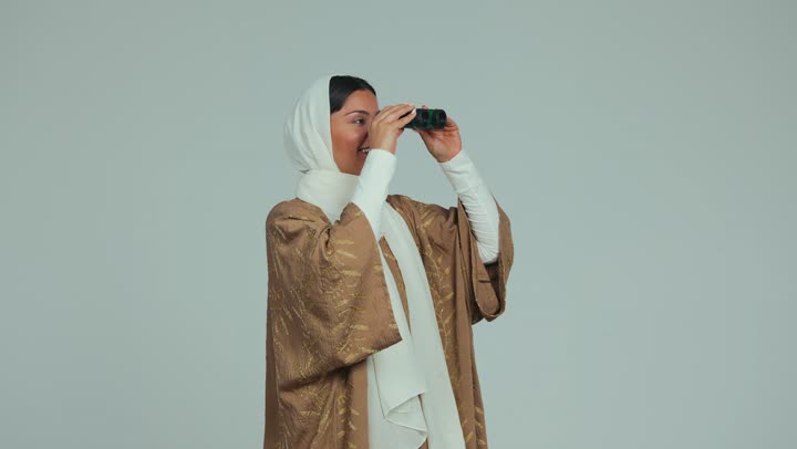 Referring to the index finger to something, monitoring and watching distant things, future vision and defining goals and aspirations, an Arab Gulf Gulf woman veiled smiling in a cloak holding her hands a binoculars binoculars and looking at the gestures of pleasure, white background