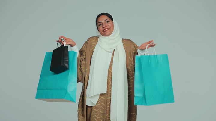 الاستفادة من العروض والتخفيضات الموسمية، الاستمتاع باقتناء المستلزمات والهدايا، أثر التسوق الإيجابي على المرأة،  سيدة عربية خليجية اماراتية محجبة مبتسمة ترتدي عباءة تحمل أكياس ورقية ملونة بإيماءات المتعة والسرور والسعادة، خلفية بيضاء