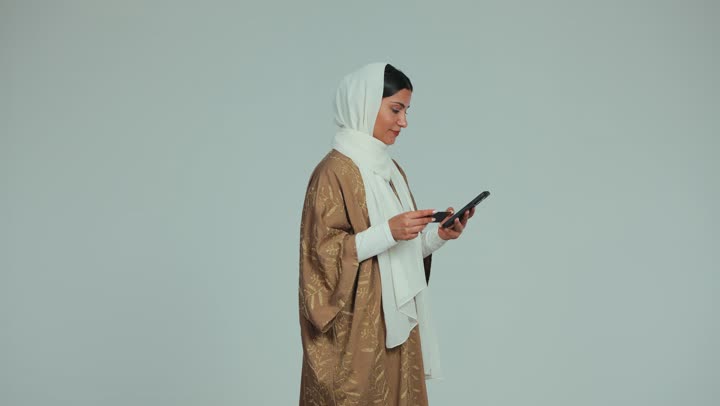 Using a modern and advanced technical device, the ability to purchase and pay remotely through programs and applications via mobile, a smiling veiled Emirati Gulf Arab woman wearing an abaya, holding a mobile phone and a bank credit card for online shopping, white background
