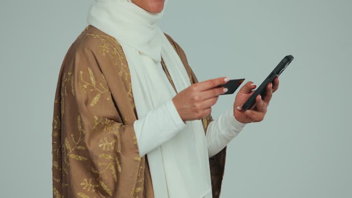 إمكانية الشراء والدفع عن بعد من خلال برامج وتطبيقات عبر الجوال، تصوير مقرب لسيدة عربية خليجية إماراتية محجبة مبتسمة ترتدي عباءة تحمل بيدها الهاتف المحمول وبطاقة الإئتمان البنكية للتسوق الالكتروني، استخدام جهاز تقني حديث ومتطور، خلفية بيضاء