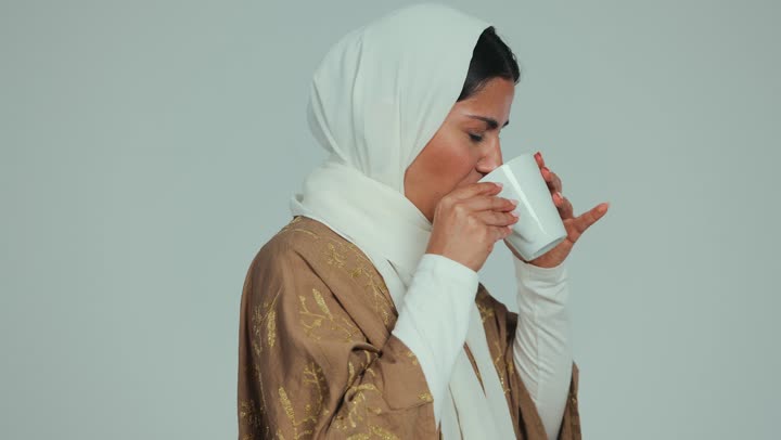 التلذذ والاستمتاع بتناول المشروب الساخن في الجو الشتوي البارد، ضرورة الإكثار من السوائل في فصل الشتاء، إيماءات تدل على البرد الشديد، الشعور بالصقيع والقشعريرة والتجمد، تصوير مقرب لسيدة عربية خليجية اماراتية محجبة ترتدي عباءة تقوم بشرب كوب من القهوة، خلفية بيضاء