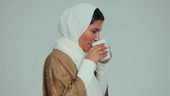 Enjoying and savoring a hot drink in the cold winter weather, the necessity of drinking plenty of fluids in the winter, gestures indicating extreme cold, feeling frostbite, chills and freezing, a close-up of a veiled Emirati Gulf Arab woman wearing an abaya drinking a cup of coffee, white background