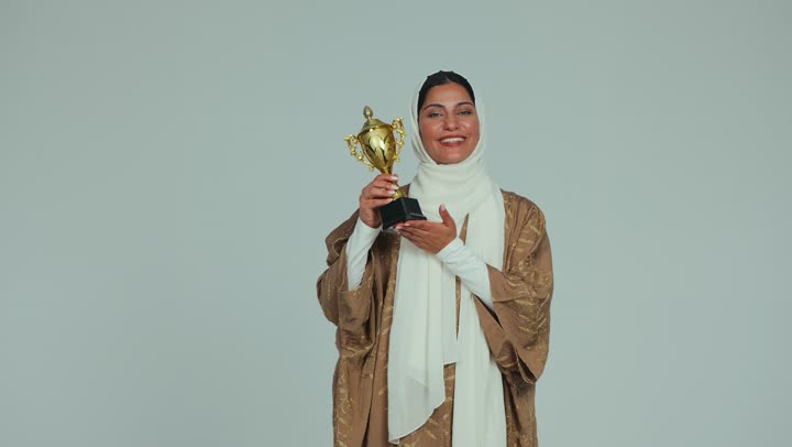 أنشطة ترفيهية ممتعة، النظر إلى الكاميرا بإيماءات الفرح والسعادة لفوز الفريق المفضل، تشجيع الأنديه والمنتخبات والفرق، امرأة عربية اماراتيه خليجية محجبة مبتسمة ترتدي العباءه تمسك بيدها الكأس الذهبي، خلفية بيضاء