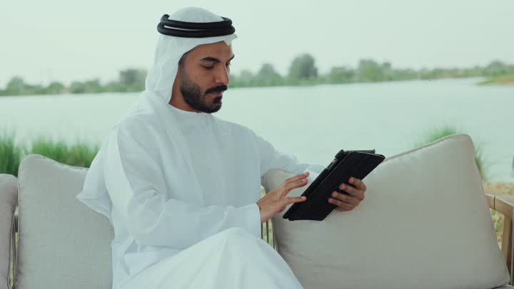 الدردشه عبر الانترنت ، تصفح مواقع التواصل الاجتماعى ،مطاعم و كافيهات في الإمارات العربية المتحدة، تصوير لرجل عربي خليجي اماراتي مبتسم يرتدي الكندورة والغترة البيضاء يجلس على الأريكة في إحدى المقاهي الراقية يمسك بيده فنجان القهوه ، الاستمتاع بالاجواء الهادئة خارج المنزل