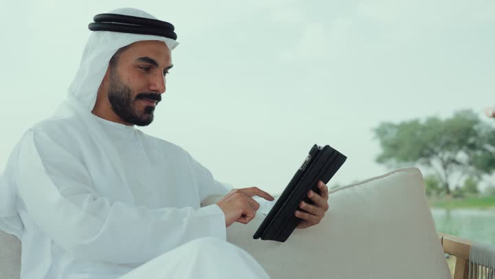 مطاعم و كافيهات في الإمارات العربية المتحدة، اجراء مكالمة فيديو مع الاهل والاقارب ، تصوير لرجل عربي خليجي اماراتي مبتسم يرتدي الكندورة والغترة البيضاء يجلس على الأريكة في إحدى المقاهي الراقية يمسك بيده فنجان القهوه ،الاستمتاع بشرب المشروبات الساخنه ، تصفح مواقع التواصل الاجتماعى ، الاستمتاع بالاجواء الهادئة خارج المنزل