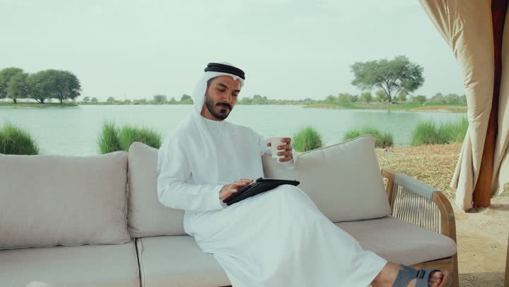 تصوير لرجل عربي خليجي اماراتي مبتسم يرتدي الكندورة والغترة البيضاء يجلس على الأريكة في إحدى المقاهي الراقية يمسك بيده فنجان القهوه ،الاستمتاع بشرب المشروبات الساخنه ، مطاعم و كافيهات في الإمارات العربية المتحدة، تصفح مواقع التواصل الاجتماعى ، الاستمتاع بالاجواء الهادئة خارج المنزل، اجراء مكالمة فيديو مع الاهل والاقارب