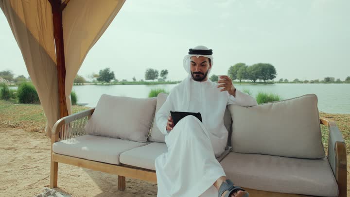 الاستمتاع بشرب المشروبات الساخنه ، مطاعم و كافيهات في الإمارات العربية المتحدة، تصفح مواقع التواصل الاجتماعى ، الاستمتاع بالاجواء الهادئة خارج المنزل، تصوير لرجل عربي خليجي اماراتي مبتسم يرتدي الكندورة والغترة البيضاء يجلس على الأريكة في إحدى المقاهي الراقية يمسك بيده فنجان القهوه ، اجراء مكالمة فيديو مع الاهل والاقارب