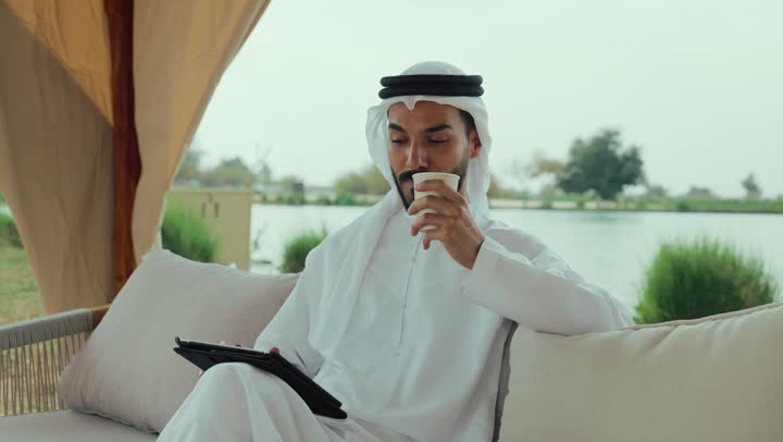 تصفح مواقع التواصل الاجتماعى ، مطاعم و كافيهات في الإمارات العربية المتحدة، الاستمتاع بالاجواء الهادئة خارج المنزل، تصوير لرجل عربي خليجي اماراتي مبتسم يرتدي الكندورة والغترة البيضاء يجلس على الأريكة في إحدى المقاهي الراقية يمسك بيده فنجان القهوه ، اجراء مكالمة فيديو مع الاهل والاقارب