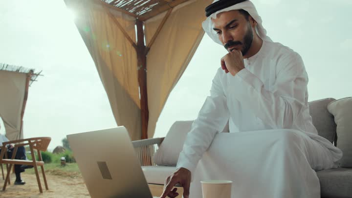الاستمتاع بالاجواء الهادئة خارج المنزل ، ايماءات الاندماج والتركيز ، تصوير لرجل عربي خليجي اماراتي يرتدي الكندورة والغترة البيضاء يجلس على الأريكة في إحدى المقاهي الراقية يقوم بتصفح الانترنت، اداره المشاريع عن بعد ، استخدام جهاز الحاسوب المحمول لانجاز المشاريع في المقهي، استخدام التكنولوجيا والتقنيات الحديثه