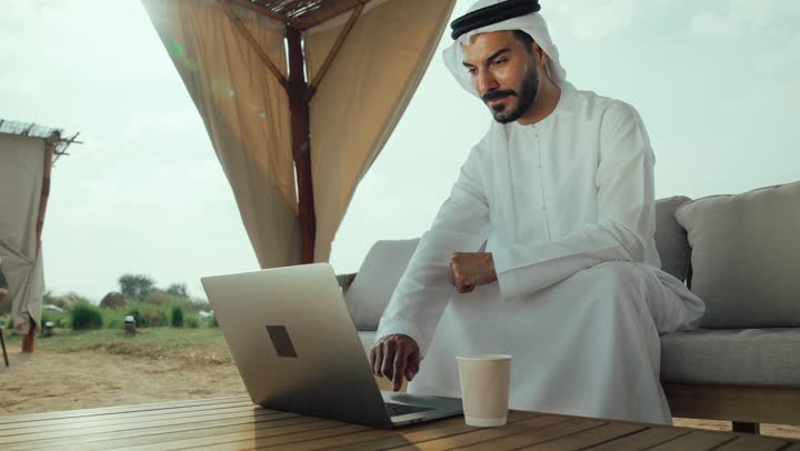 ايماءات الاندماج والتركيز ، تصوير لرجل عربي خليجي اماراتي يرتدي الكندورة والغترة البيضاء يجلس على الأريكة في إحدى المقاهي الراقية يقوم بتصفح الانترنت، اداره المشاريع عن بعد ، استخدام جهاز الحاسوب المحمول لانجاز المشاريع في المقهي، استخدام التكنولوجيا والتقنيات الحديثه ، الاستمتاع بالاجواء الهادئة خارج المنزل