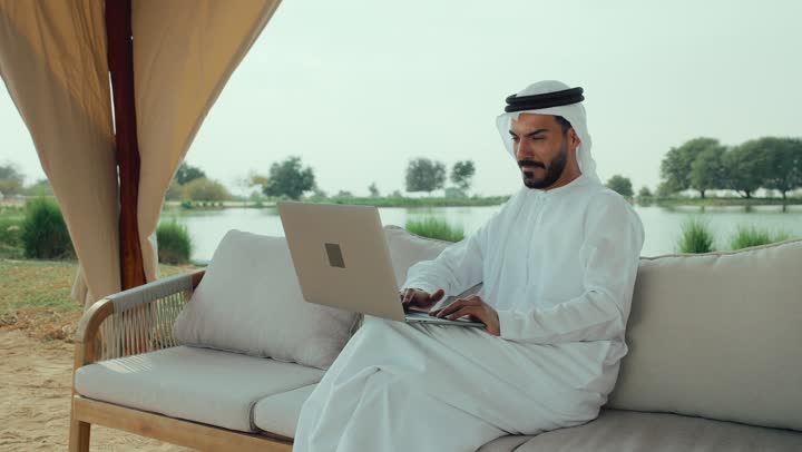 تصوير لرجل عربي خليجي اماراتي يرتدي الكندورة والغترة البيضاء يجلس على الأريكة في إحدى المقاهي الراقية يقوم بتصفح الانترنت، اداره المشاريع عن بعد ، استخدام جهاز الحاسوب المحمول لانجاز المشاريع في المقهي، استخدام التكنولوجيا والتقنيات الحديثه ، الاستمتاع بالاجواء الهادئة خارج المنزل