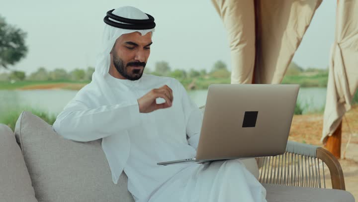 استخدام جهاز الحاسوب المحمول لانجاز المشاريع في المقهي، استخدام التكنولوجيا والتقنيات الحديثه ، الاستمتاع بالاجواء الهادئة خارج المنزل، انجاز واداره المشاريع عن بعد ، رجل عربي خليجي اماراتي يرتدي الكندورة والغترة البيضاء يجلس على الأريكة في إحدى المقاهي الراقية يقوم بتصفح الانترنت