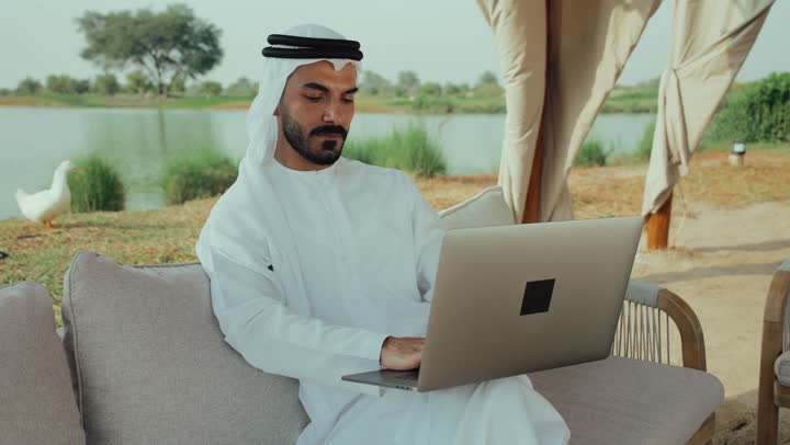 استخدام التكنولوجيا والتقنيات الحديثه ، الاستمتاع بالاجواء الهادئة خارج المنزل، انجاز واداره المشاريع عن بعد ، استخدام جهاز الحاسوب المحمول لانجاز المشاريع في المقهي، رجل عربي خليجي اماراتي يرتدي الكندورة والغترة البيضاء يجلس على الأريكة في إحدى المقاهي الراقية يقوم بتصفح الانترنت