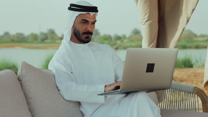 الاستمتاع بالاجواء الهادئة خارج المنزل، انجاز واداره المشاريع عن بعد ، استخدام جهاز الحاسوب المحمول لانجاز المشاريع في المقهي، رجل عربي خليجي اماراتي يرتدي الكندورة والغترة البيضاء يجلس على الأريكة في إحدى المقاهي الراقية يقوم بتصفح الانترنت