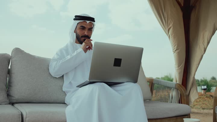 استخدام جهاز الحاسوب المحمول لانجاز المشاريع في المقهي، انجاز واداره المشاريع عن بعد ، رجل عربي خليجي اماراتي يرتدي الكندورة والغترة البيضاء يجلس على الأريكة في إحدى المقاهي الراقية يقوم بتصفح الانترنت، الاستمتاع بالاجواء الهادئة خارج المنزل
