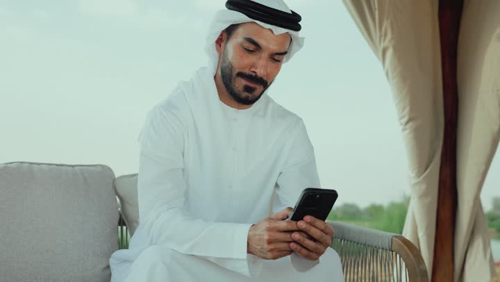تصفح مواقع التواصل الاجتماعي عن طريق الانترنت ، تصوير لرجل عربي خليجي اماراتي يرتدي الكندورة والغترة البيضاء يجلس على الأريكة في إحدى المقاهي الراقية يقوم بتصفح الجوال، الاستمتاع بالاجواء الهادئة خارج المنزل، استخدام الهاتف المحمول ، مطاعم و كافيهات في الإمارات العربية المتحدة