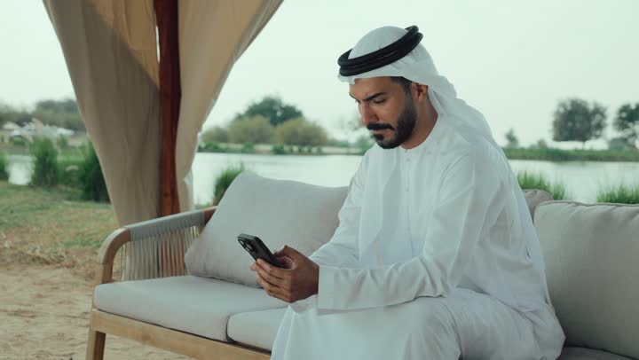 تصوير لرجل عربي خليجي اماراتي يرتدي الكندورة والغترة البيضاء يجلس على الأريكة في إحدى المقاهي الراقية يقوم بتصفح الجوال، الاستمتاع بالاجواء الهادئة خارج المنزل، استخدام الهاتف المحمول ، تصفح مواقع التواصل الاجتماعي عن طريق الانترنت ، مطاعم و كافيهات في الإمارات العربية المتحدة