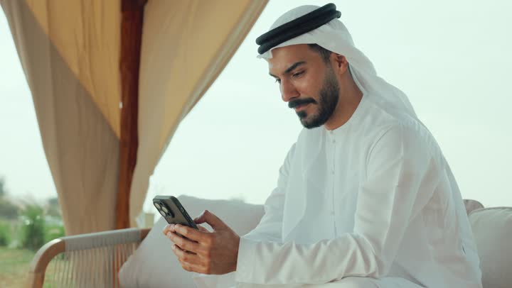 الاستمتاع بالاجواء الهادئة خارج المنزل، استخدام الهاتف المحمول ، تصوير لرجل عربي خليجي اماراتي يرتدي الكندورة والغترة البيضاء يجلس على الأريكة في إحدى المقاهي الراقية يقوم بتصفح الجوال،  تصفح مواقع التواصل الاجتماعي عن طريق الانترنت ، مطاعم و كافيهات في الإمارات العربية المتحدة
