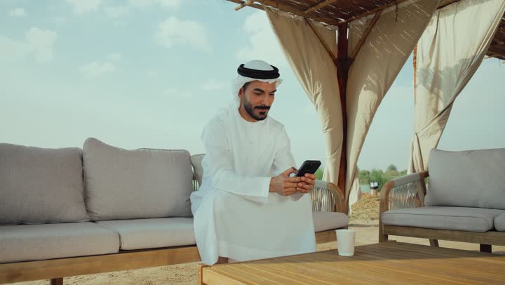 استخدام الهاتف المحمول ، تصوير لرجل عربي خليجي اماراتي يرتدي الكندورة والغترة البيضاء يجلس على الأريكة في إحدى المقاهي الراقية يقوم بتصفح الجوال،  تصفح مواقع التواصل الاجتماعي عن طريق الانترنت ، الاستمتاع بالاجواء الهادئة خارج المنزل، مطاعم و كافيهات في الإمارات العربية المتحدة