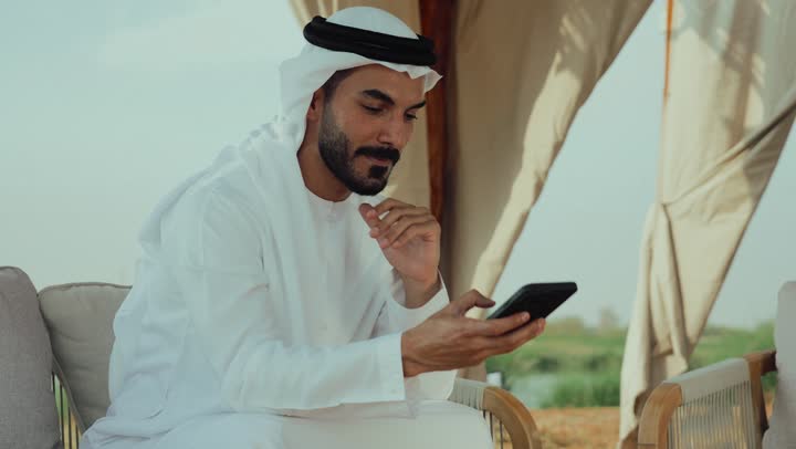 الاطمئنان على الاهل والاصدقاء ، تصوير لرجل عربي خليجي اماراتي يرتدي الكندورة والغترة البيضاء يجلس على الأريكة في إحدى المقاهي الراقية يقوم بتصفح الجوال، تصفح مواقع التواصل الاجتماعي عن طريق الانترنت ، الاستمتاع بالاجواء الهادئة خارج المنزل، مطاعم و كافيهات في الإمارات العربية المتحدة