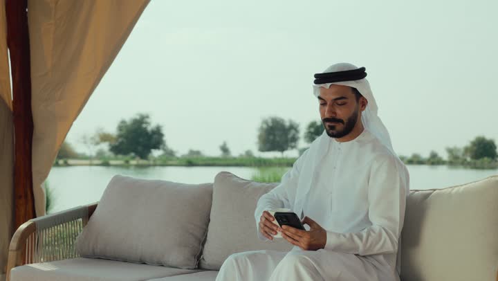 االدردشه عبر الانترنت ، تصوير لرجل عربي خليجي اماراتي يرتدي الكندورة والغترة البيضاء يجلس على الأريكة في إحدى المقاهي الراقية يقوم بتصفح الجوال،  تصفح مواقع التواصل الاجتماعي عن طريق الانترنت ، الاستمتاع بالاجواء الهادئة خارج المنزل، مطاعم و كافيهات في الإمارات العربية المتحدة