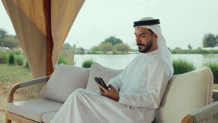 اجراء مكالمات الفيديو ، تصوير لرجل عربي خليجي اماراتي يرتدي الكندورة والغترة البيضاء يجلس على الأريكة في إحدى المقاهي الراقية يقوم بتصفح الجوال، تصفح مواقع التواصل الاجتماعي عن طريق الانترنت ، الاستمتاع بالاجواء الهادئة خارج المنزل، مطاعم و كافيهات في الإمارات العربية المتحدة