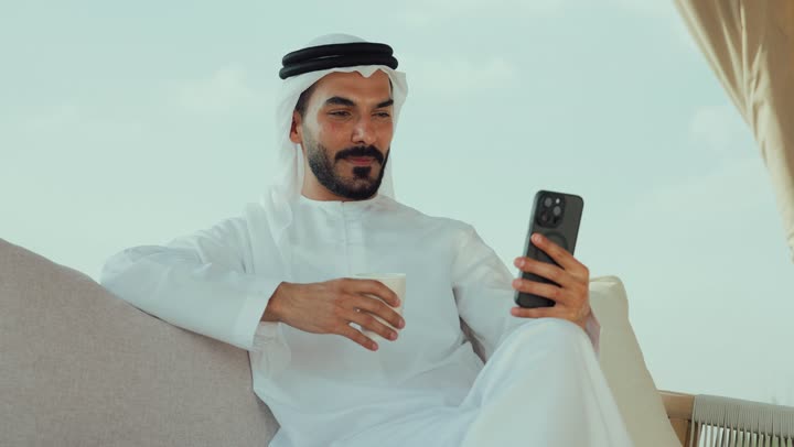اجراء بث مباشر ، تصوير لرجل عربي خليجي اماراتي يرتدي الكندورة والغترة البيضاء يجلس على الأريكة في إحدى المقاهي الراقية يقوم بتصفح الجوال، تصفح مواقع التواصل الاجتماعي عن طريق الانترنت ، الاستمتاع بالاجواء الهادئة خارج المنزل، مطاعم و كافيهات في الإمارات العربية المتحدة