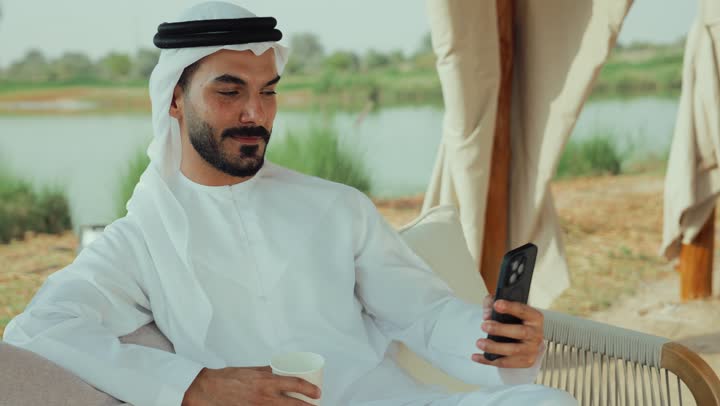 تصوير لرجل عربي خليجي اماراتي يرتدي الكندورة والغترة البيضاء يجلس على الأريكة في إحدى المقاهي الراقية يقوم بتصفح الجوال،  تصفح مواقع التواصل الاجتماعي عن طريق الانترنت ، الاستمتاع بالاجواء الهادئة خارج المنزل، مطاعم و كافيهات في الإمارات العربية المتحدة