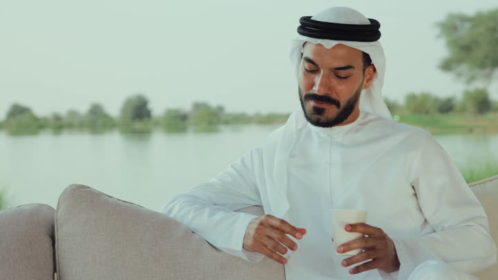 تصفح مواقع التواصل الاجتماعي عن طريق الانترنت ، الاستمتاع بالاجواء الهادئة خارج المنزل، مطاعم و كافيهات في الإمارات العربية المتحدة، تصوير لرجل عربي خليجي اماراتي يرتدي الكندورة والغترة البيضاء يجلس على الأريكة في إحدى المقاهي الراقية يقوم بتصفح الجوال