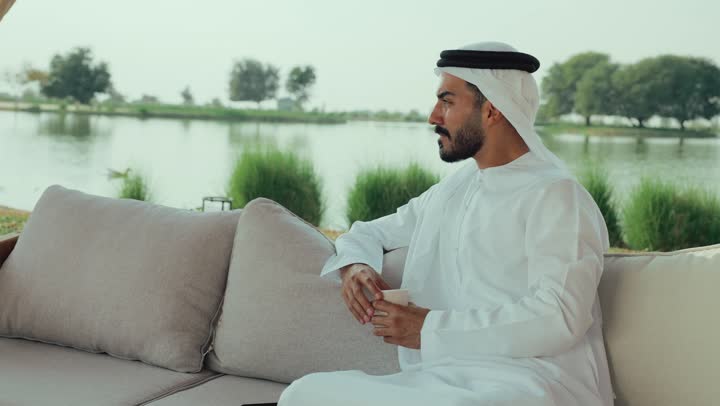 الاستمتاع بالاجواء الهادئة خارج المنزل، مطاعم و كافيهات في الإمارات العربية المتحدة، تصوير لرجل عربي خليجي اماراتي يرتدي الكندورة والغترة البيضاء يجلس على الأريكة في إحدى المقاهي الراقية يقوم بتصفح الجوال ، تصفح مواقع التواصل الاجتماعي عن طريق الانترنت