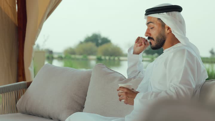 مطاعم و كافيهات في الإمارات العربية المتحدة، الاستمتاع بالاجواء الهادئة خارج المنزل، تصوير لرجل عربي خليجي اماراتي يرتدي الكندورة والغترة البيضاء يجلس على الأريكة في إحدى المقاهي الراقية يقوم بتصفح الجوال ، تصفح مواقع التواصل الاجتماعي عن طريق الانترنت