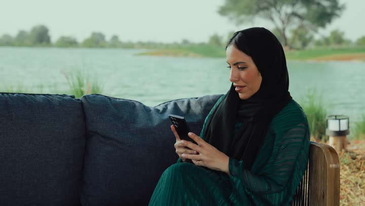 تبادل الأحاديث المسليه عبر الجوال، الدردشة والتواصل الاجتماعي، استخدام التكنولوجيا الحديثه ، سيده سعودية تقوم باستخدام الجوال ،قضاء وقت ممتع فى الخارج ، ايماءات السعاده والسرور، تصوير لامراة عربية خليجية اماراتية محجبة ترتدي العباءة تجلس على الأريكة في إحدى المقاهي الراقية تستمع بلحظات الهدوء والراحة