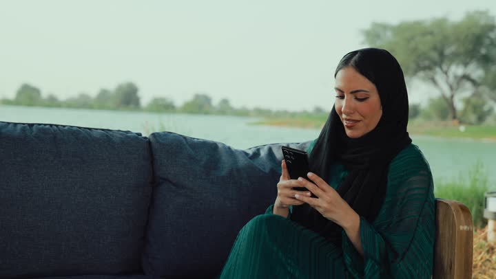 الدردشة  والتواصل الاجتماعي، استخدام التكنولوجيا الحديثه ، سيده سعودية تقوم باستخدام الجوال ،قضاء وقت ممتع فى الخارج ، ايماءات السعاده والسرور، تصوير لامراة عربية خليجية اماراتية محجبة ترتدي العباءة تجلس على الأريكة في إحدى المقاهي الراقية تستمع بلحظات الهدوء والراحة