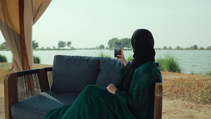 توثيق اللحظات السعيده ، سيده سعودية تقوم باستخدام التقنيات الحديثه ،قضاء وقت ممتع عبر الجوال مع الأصحاب، ايماءات السعاده والسرور، تصوير لامراة عربية خليجية اماراتية محجبة ترتدي العباءة تجلس على الأريكة في إحدى المقاهي الراقية تستمع بلحظات الهدوء والراحة