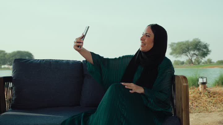 تبادل الأحاديث المسليه عبر الجوال، سيده سعودية تقوم باجراء مكالمة فيديو ،قضاء وقت ممتع عبر الجوال مع الأصحاب، ايماءات السعاده والسرور، تصوير لامراة عربية خليجية اماراتية محجبة ترتدي العباءة تجلس على الأريكة في إحدى المقاهي الراقية تستمع بلحظات الهدوء والراحة