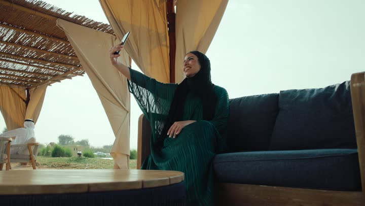 التواصل و الاطمئنان على الأصدقاء والأقارب، سيده سعودية تقوم باجراء مكالمة فيديو ،قضاء وقت ممتع عبر الجوال مع الأصحاب، اجراء بث مباشر ، ايماءات السعاده والسرور، تصوير لامراة عربية خليجية اماراتية محجبة ترتدي العباءة تجلس على الأريكة في إحدى المقاهي الراقية تستمع بلحظات الهدوء والراحة