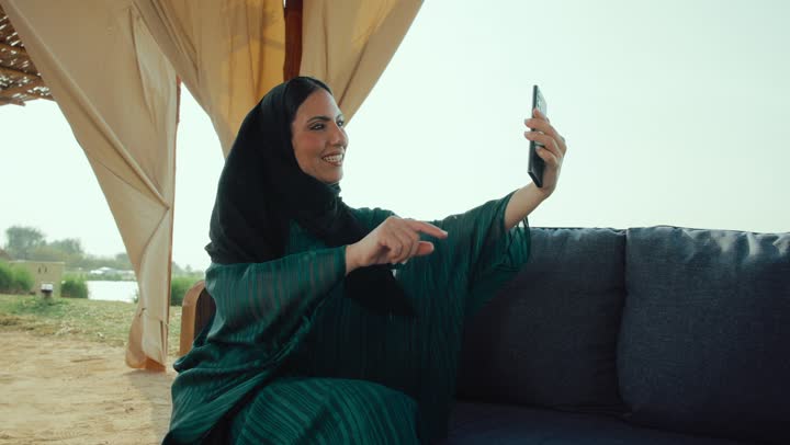 قضاء وقت ممتع عبر الجوال مع الأصحاب، اجراء بث مباشر ، ايماءات السعاده والسرور، تصوير لامراة عربية خليجية اماراتية محجبة ترتدي العباءة تجلس على الأريكة في إحدى المقاهي الراقية تستمع بلحظات الهدوء والراحة ، سيده سعودية تقوم باجراء مكالمة فيديو