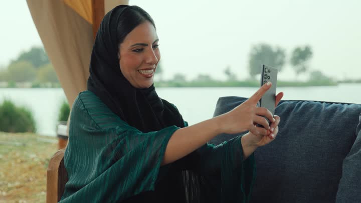 اجراء بث مباشر ، ايماءات السعاده و السرور، قضاء وقت ممتع عبر الجوال مع الأصحاب، تصوير لامراة عربية خليجية اماراتية محجبة ترتدي العباءة تجلس على الأريكة في إحدى المقاهي الراقية تستمع بلحظات الهدوء والراحة ، سيده سعودية تقوم باجراء مكالمة فيديو