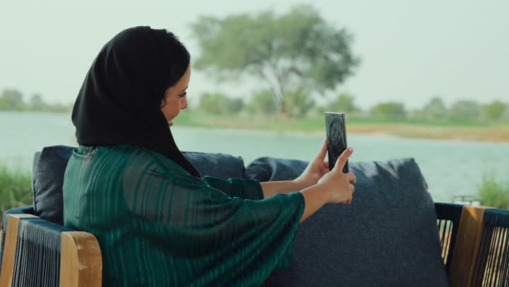 ايماءات السعاده و السرور، قضاء وقت ممتع عبر الجوال مع الأصحاب، احتساء المشروب المفضل، تصوير لامراة عربية خليجية اماراتية محجبة ترتدي العباءة تجلس على الأريكة في إحدى المقاهي الراقية تستمع بلحظات الهدوء والراحة ، سيده سعودية تقوم باجراء مكالمة فيديو